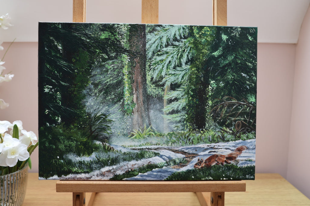 Painting of a forest scene with a red squirrel and snow on the ground on an easel.