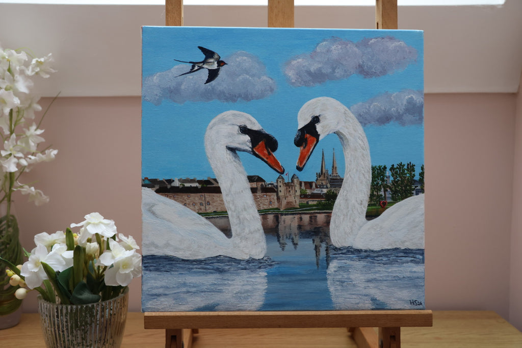 Painting of two swans on Lough Erne with the townscape of Enniskillen in the background, displayed on an easel.