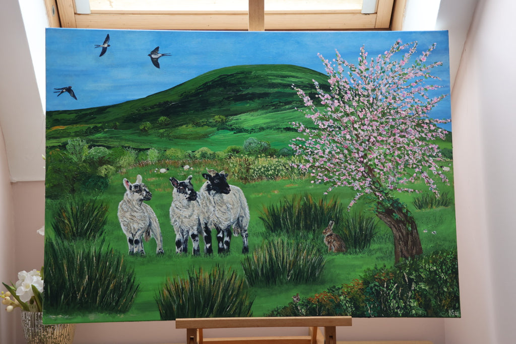 Painting of three lambs in a pastoral scene with hills and flowers on an easel.