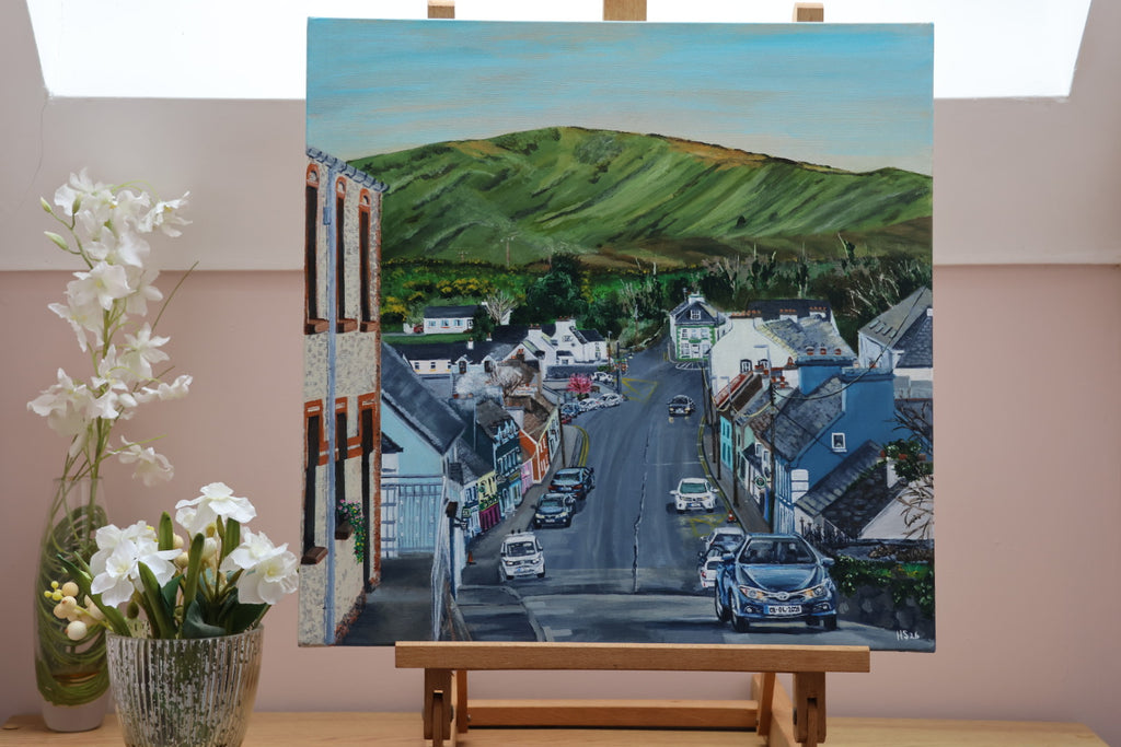Painting of the town street of Ardara with cars and green hills on an easel against a light pink wall.